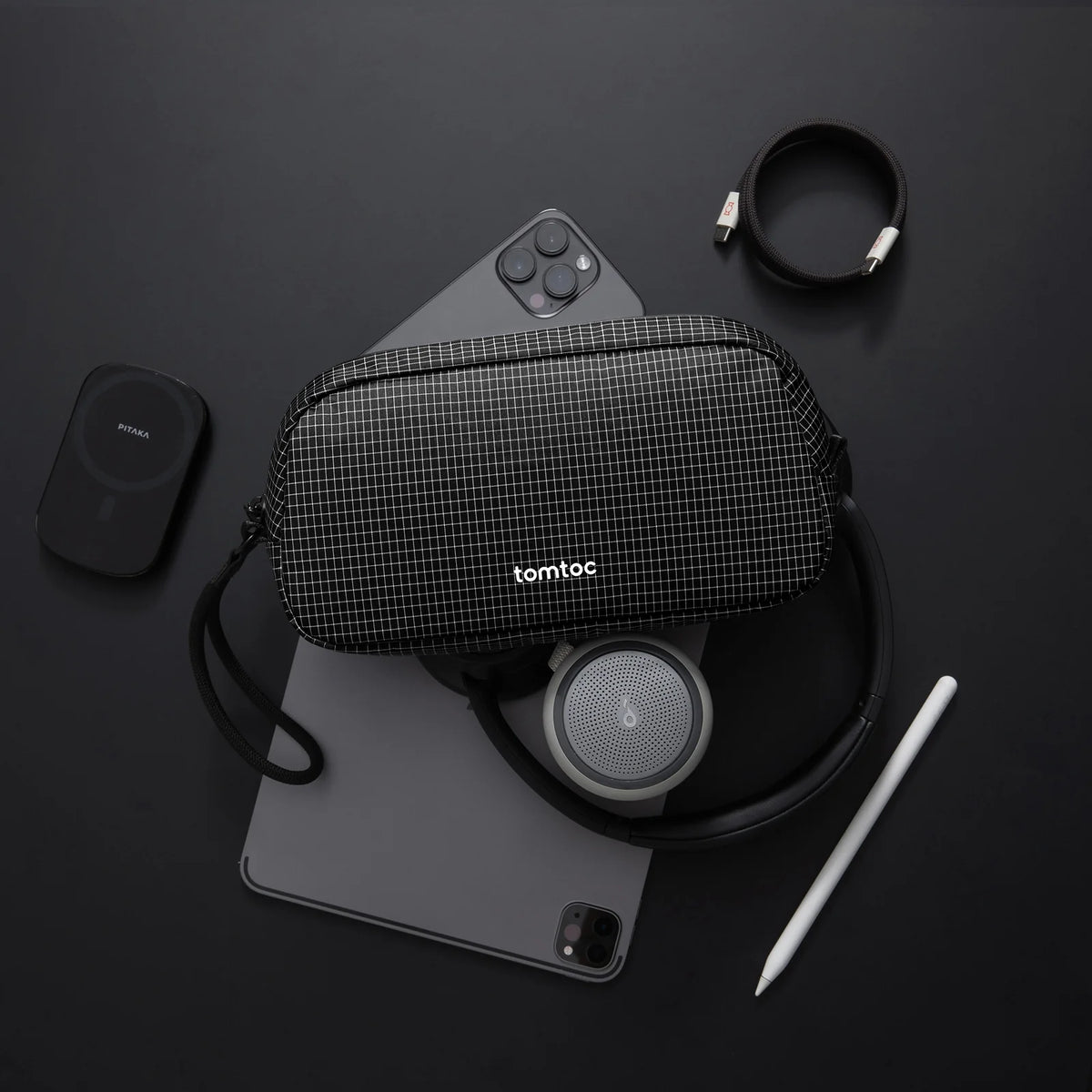 Black grid-patterned pouch with 'tomtoc' branding on a black surface with electronic items.