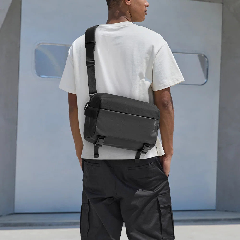 A man wearing a white t-shirt and black cargo pants, viewed from behind, carrying a darktomtoc T21L1D1 Business laptop sling bag on his back.