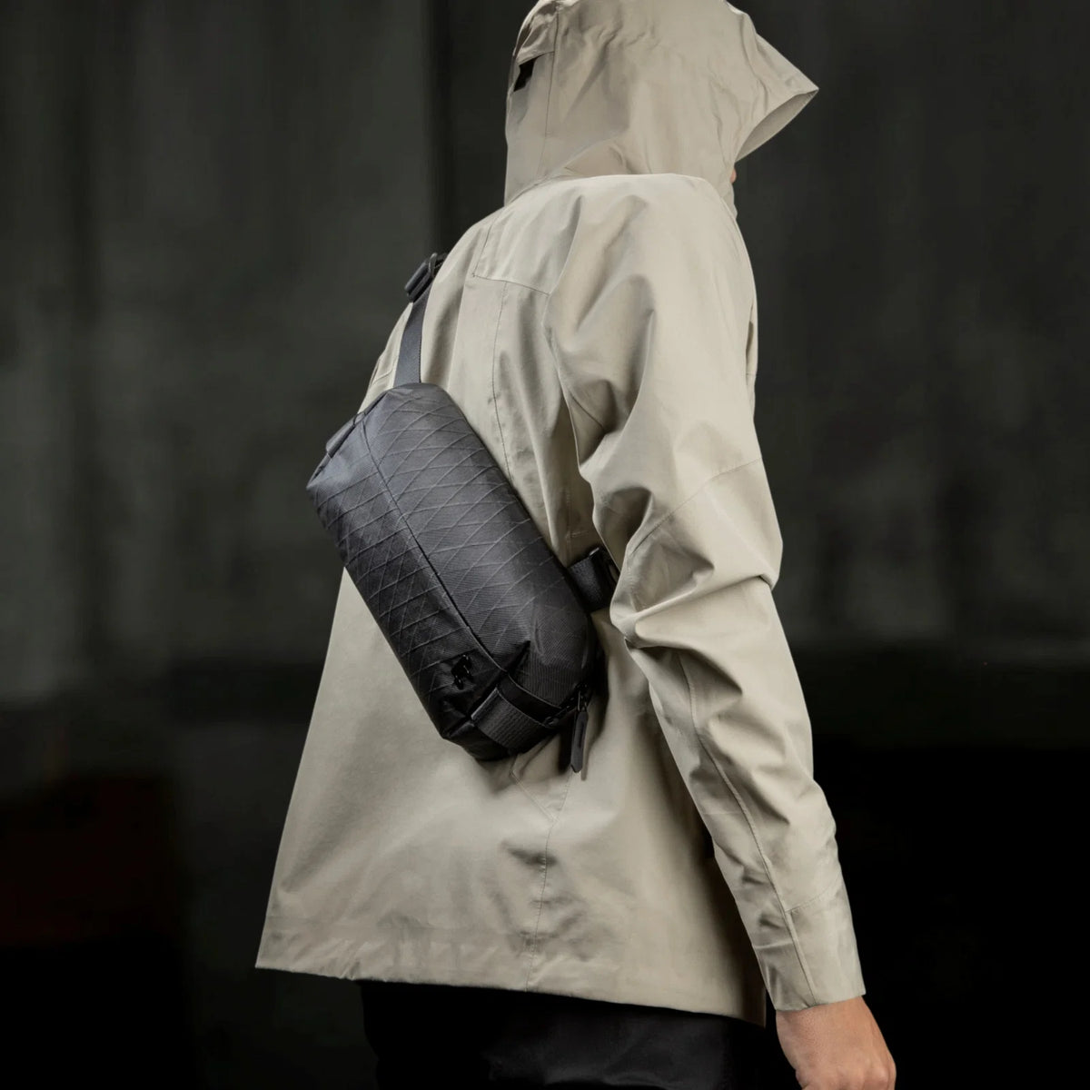 A person wearing a light beige hooded jacket from the back, carrying a tomtoc X-Pac Sling Bag Small (3L) T21S1D2 on their shoulder against a dark gray background.