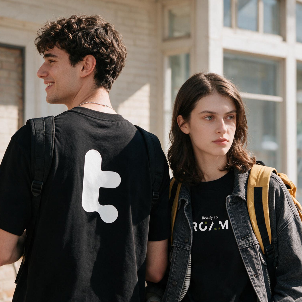 Two young adults standing outdoors in front of a building, each wearing a tomtoc Core Unisex Roam Logo Tee. The person on the left shows the back of the shirt with a large white abstract symbol, while the person on the right shows the front chest area with white text reading 'Ready To ROAM'.