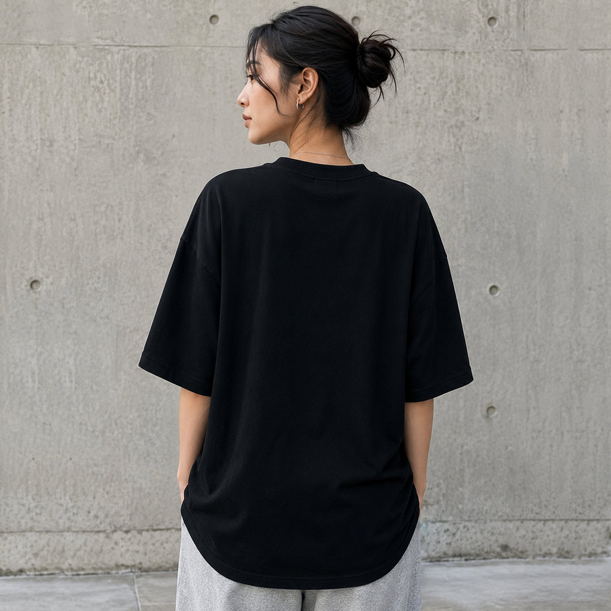 A woman viewed from behind wearing an tomtoc Signature Unisex Embroidery Tee and light grey sweatpants, standing against a textured concrete wall.