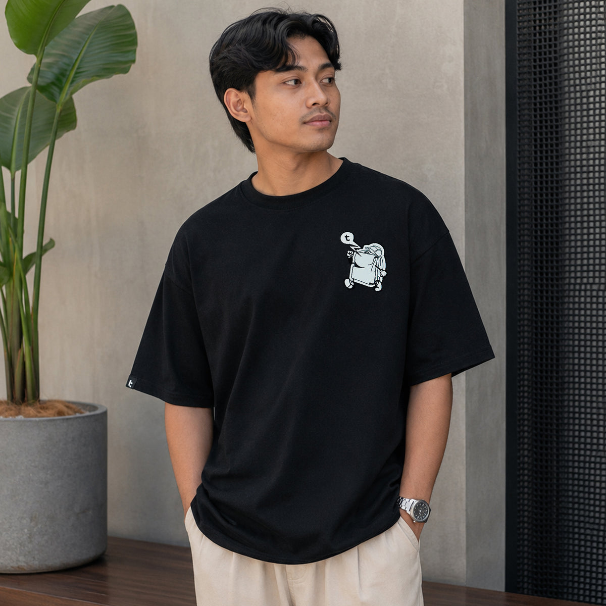 A man wearing a black tomtoc Signature Unisex Embroidery Tee with a small white cartoon graphic on the left chest, paired with beige trousers, standing in a modern indoor setting.