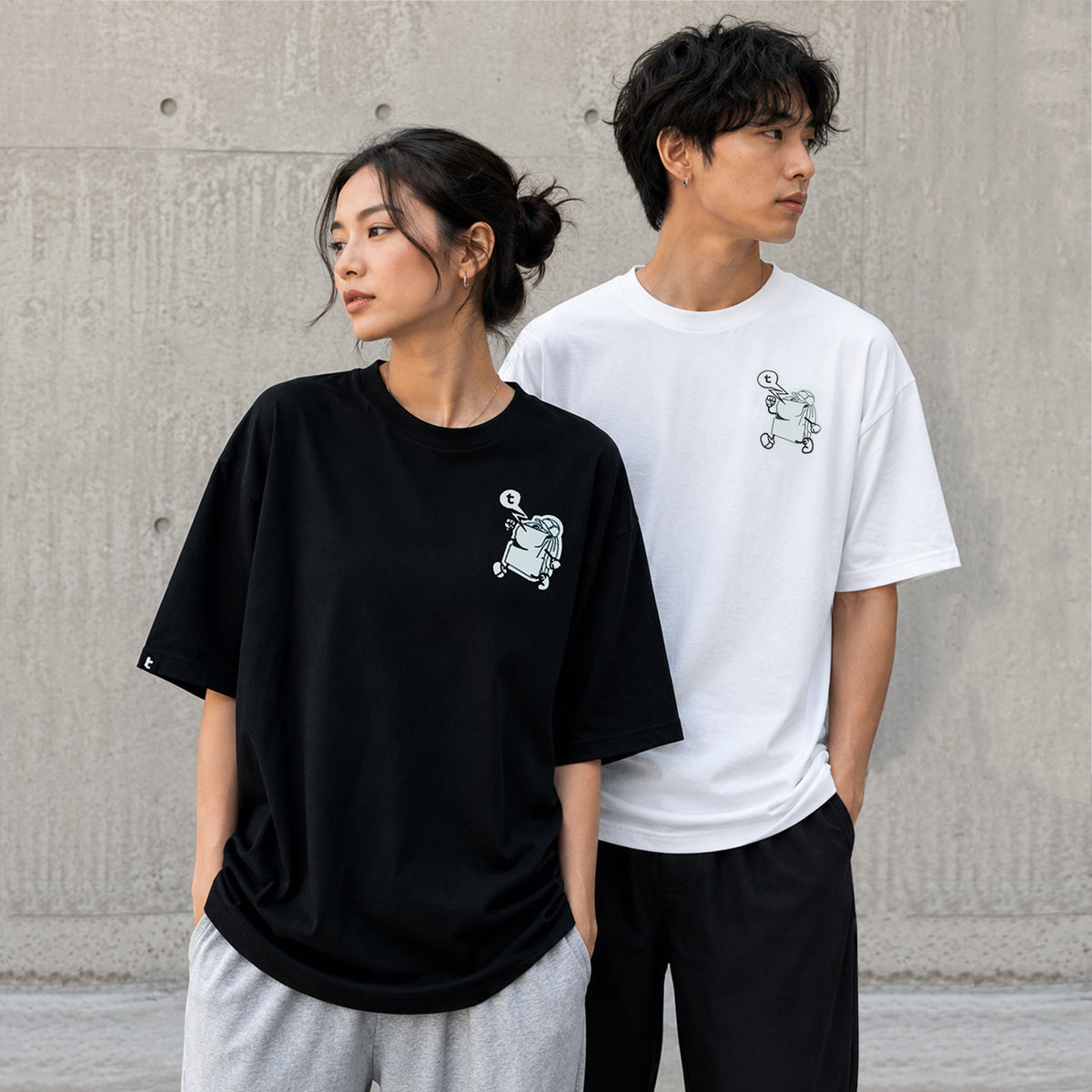 A man and a woman modeling oversized graphic T-shirts in front of a concrete wall. The woman wears a black tomtoc Signature Unisex Embroidery Tee with a small white graphic on the left chest, paired with light gray sweatpants.